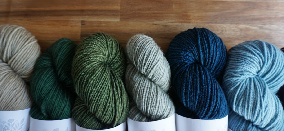 Six skeins of yarn in assorted colors arranged in a row against a wooden plank background. The yarn colors include beige, forest green, olive green, light sage green, deep navy blue, and dusty blue.